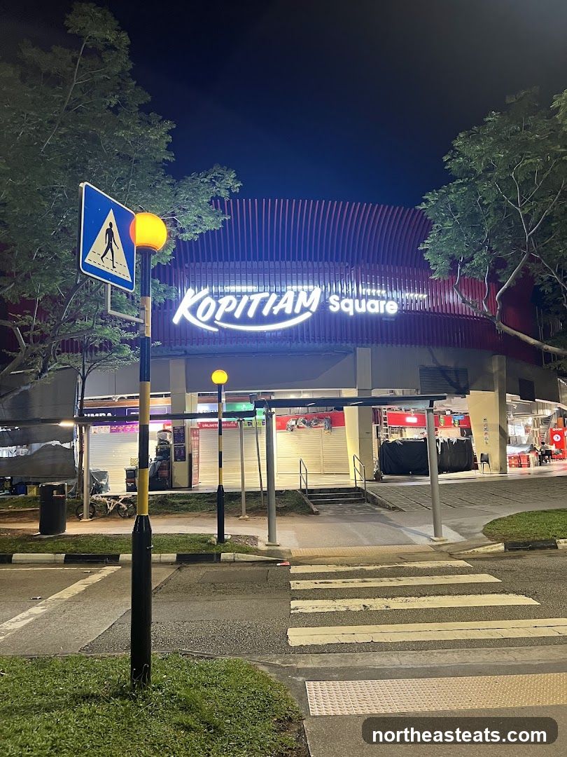 A night view of the exterior of Kopitiam Square, a building with a prominent neon sign. The building has a modern architectural design with red vertical slats on its upper facade. In the foreground, there is a pedestrian crossing and a street light with a pedestrian crossing sign. Some shops within the square appear to be closed, with their shutters down.
