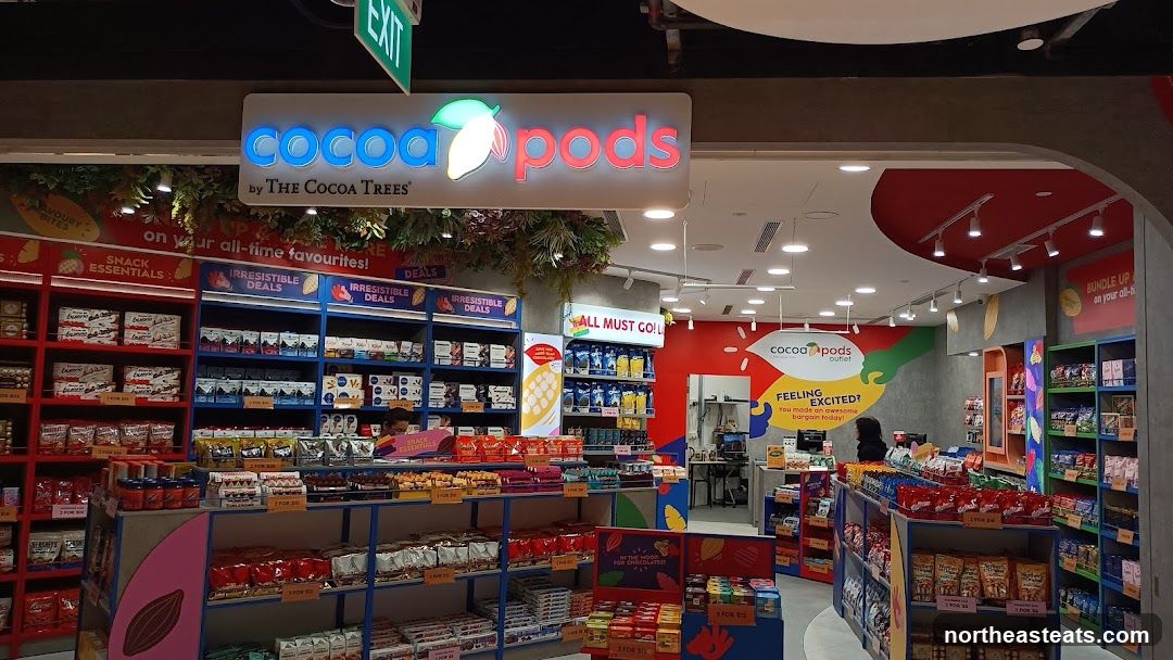 A brightly lit store interior with shelves stocked full of chocolates and snacks. The store's name, "Cocoa Pods by The Cocoa Trees," is prominently displayed above the entrance. Various promotional signs are visible, including "Irresistible Deals," "Snack Essentials," and "All Must Go!" A person is visible behind the counter.