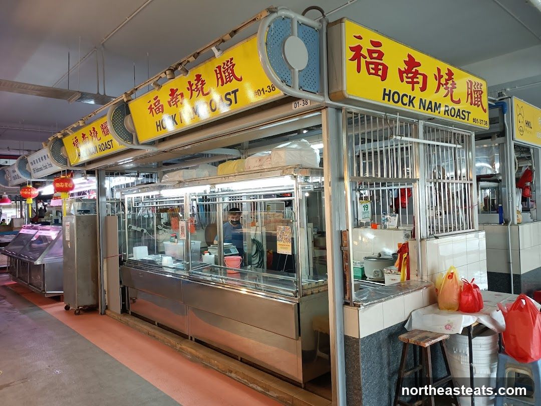 A food stall with a prominent yellow sign displaying "HOCK NAM ROAST" in both Chinese and English. The stall features a glass display case with food items and a person working behind the counter. There are also metal grates and various containers visible. The stall is located in an indoor market setting.