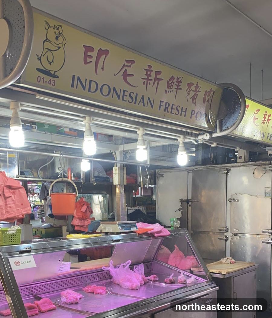 A stall with a yellow sign featuring a cartoon pig and the text "INDONESIAN FRESH PORK" in English and Chinese. Below the sign, a display case filled with raw pork cuts, some wrapped in plastic bags. In the background, there are large refrigerators and scales.