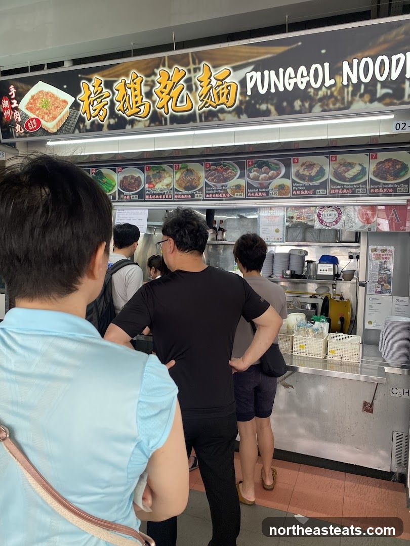 A queue of people stand in front of a food stall with a large sign above that reads "PUNGGOL NOODLES" in English and Chinese characters. Below the sign are pictures of various noodle dishes with their names and prices. The stall appears to be a hawker stall with food preparation areas visible behind the counter.