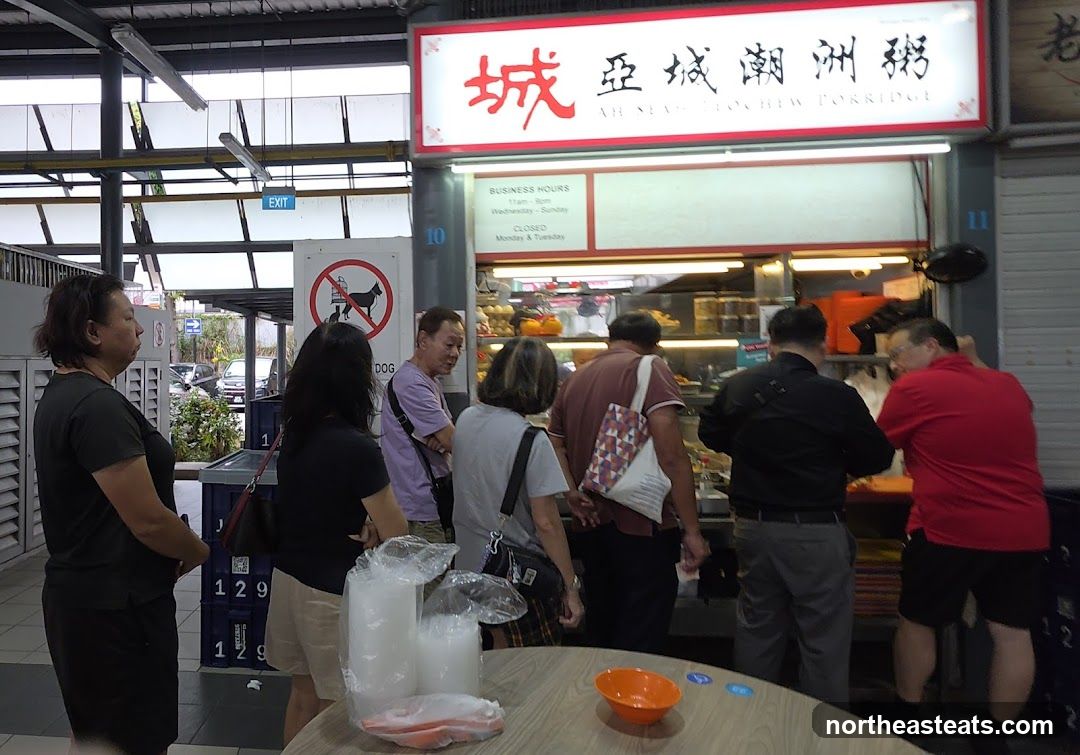 A food stall with a sign that reads "Ah Nea Rochew Porridge" in English and Chinese characters. The stall has a display of food items and customers are queuing. Business hours are posted, indicating it is open from 11 am to 8 pm, Wednesday to Sunday, and closed on Mondays and Tuesdays. There is also a "No Dogs Allowed" sign.