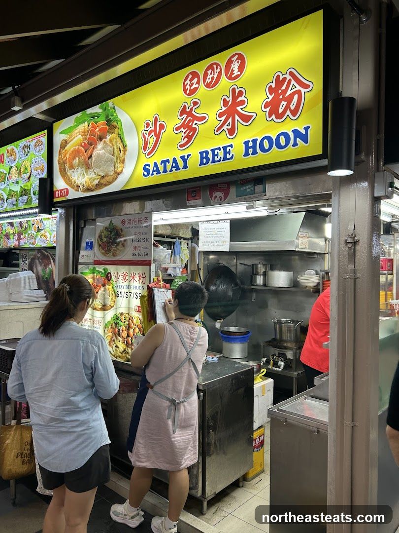 A brightly lit stall with a large yellow sign advertising "SATAY BEE HOON" in English and Chinese. Below the main sign, there are smaller menu boards displaying prices for various dishes. Two customers are standing in front of the stall, one looking at a menu and the other at a clipboard. The stall appears to be in a food court or hawker center setting, with kitchen equipment visible in the background.