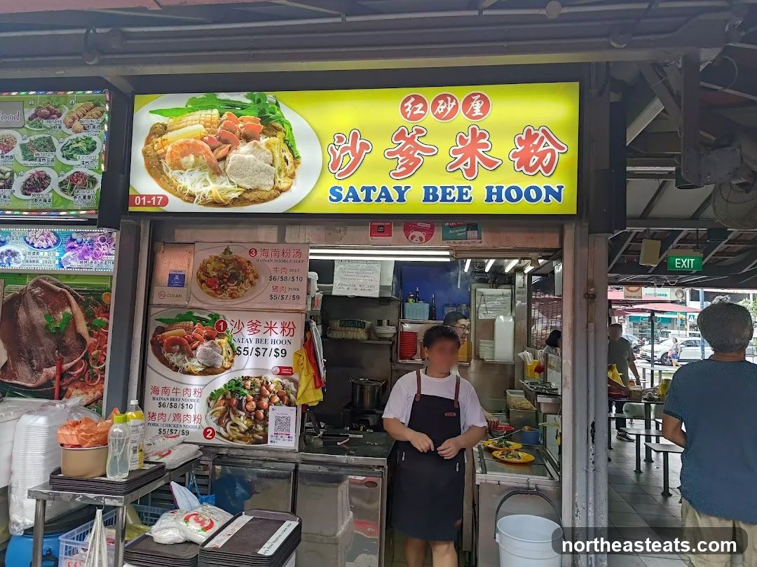 A food stall with a large yellow sign displaying "SATAY BEE HOON" in English and Chinese. Below the main sign are menu boards with pictures and prices for various noodle dishes. A person in an apron stands behind the counter. The stall appears to be part of an outdoor food court or hawker center.