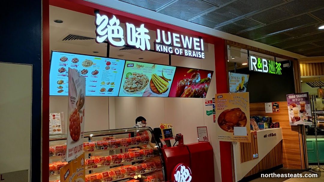 The exterior of a JUEWEI King of Braise food stall with a large digital menu board displaying various braised items. To the right, an R&B Tea stall is visible. The JUEWEI stall has a display case filled with packaged food items and a counter with a cashier. Promotional posters for "Smoked Whole Chicken" and "Taro Tango" are displayed.