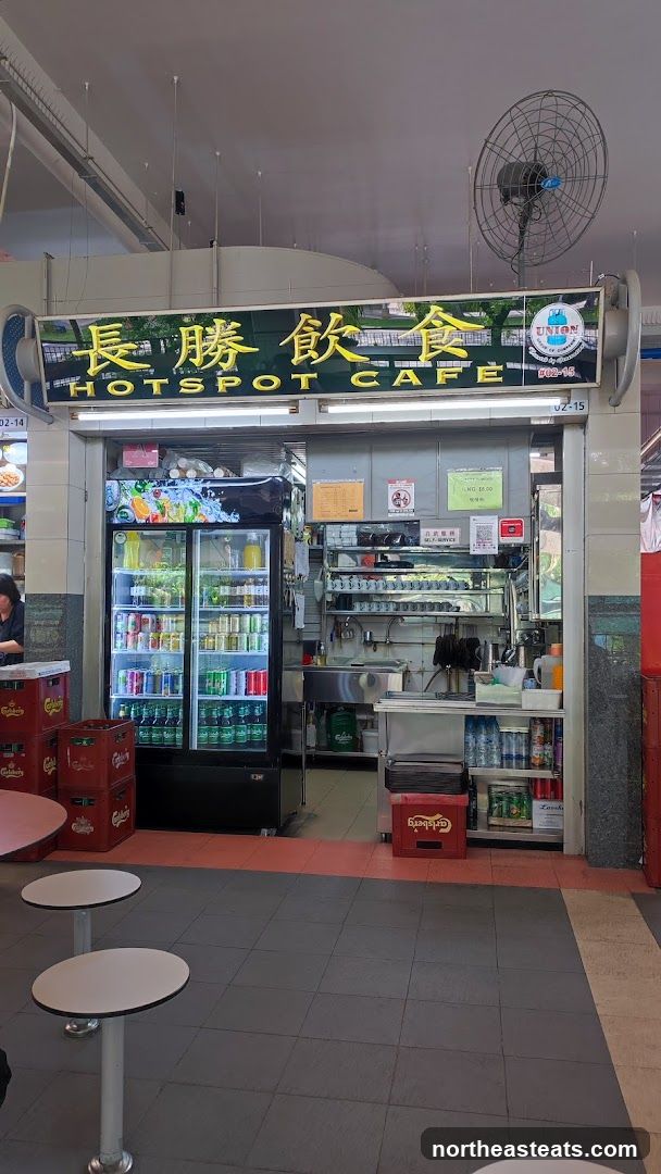 The exterior of a cafe with a prominent sign that reads "HOT SPOT CAFE" in English and Chinese characters. To the left of the entrance is a large glass-door refrigerator stocked with various beverages. The interior shows a counter area with shelves, a sink, and kitchen equipment. There are also tables and chairs visible outside the cafe.