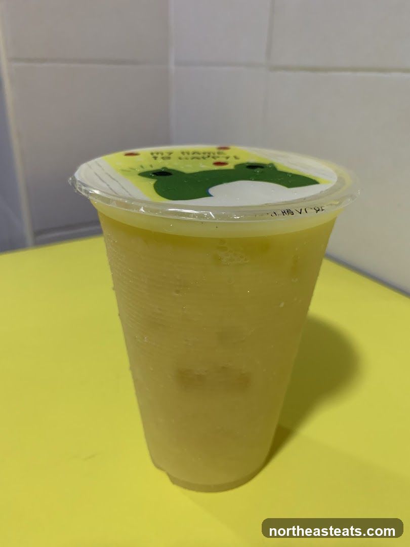 A frosted plastic cup filled with a yellow beverage and ice cubes. The cup has a clear plastic lid with a yellow sticker featuring a cartoon frog and the text "MY NAME TO HAPPY!". The cup is placed on a yellow surface, with a white tiled wall in the background.