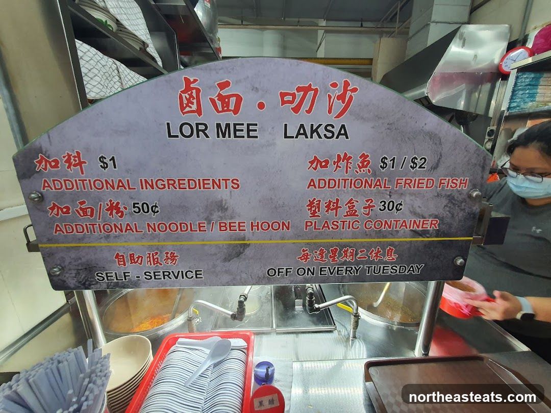A menu board displaying prices for Lor Mee and Laksa, with options for additional ingredients and fried fish.
