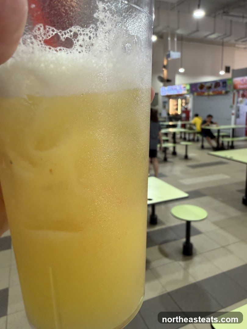 A close-up shot of a frothy yellow beverage in a clear plastic cup, held by a hand on the left. The drink appears to be a juice or smoothie with a foamy head. In the blurred background, a food court setting is visible with tables and chairs, and people walking around. Several food stalls with colorful signage are also discernible in the distance.