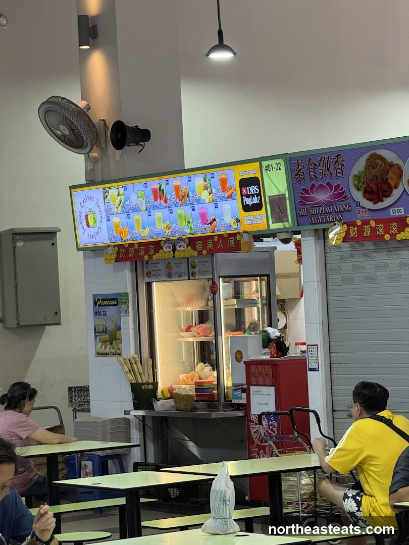 A food stall in a hawker center, featuring a brightly lit menu board displaying various fruit juices and a separate menu for vegetarian dishes. The stall also has a display fridge with fresh fruits and drinks. Customers are seated at tables in the foreground.